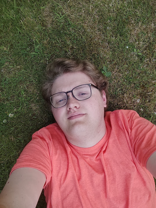 Andrew lying on the grass, wearing a t-shirt somewhere in colour between orange and pink